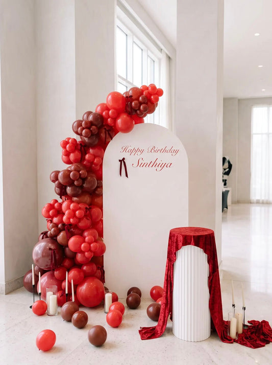White birthday backdrop with red and burgundy balloon garland, bow accents, personalized lettering, candles and a red velvet-draped display stand.