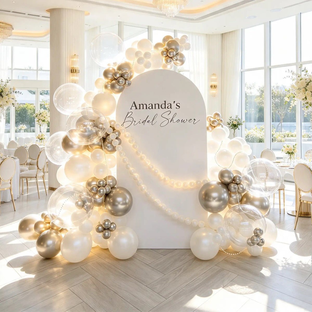White bridal shower arch with personalized lettering, ivory and champagne gold balloons, clear bubbles, and draped pearl-inspired garlands.