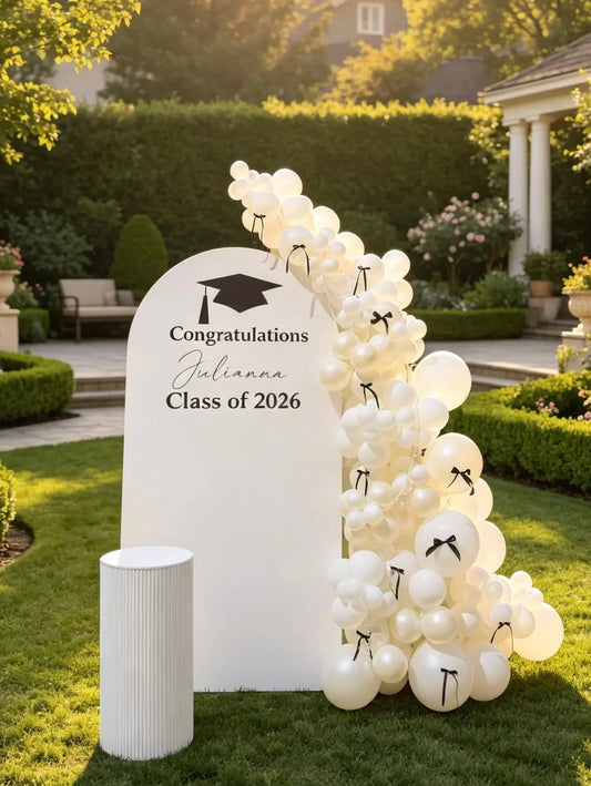 White Class of 2026 graduation backdrop with an all-white organic balloon garland, black bow accents, pearl details and a personalized graduate name.