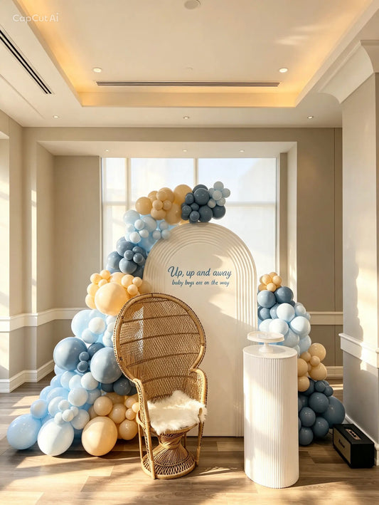 Cream baby shower arch with “Up, up and away” lettering, blue and cream balloons, a wicker peacock chair, and a white cake pedestal.
