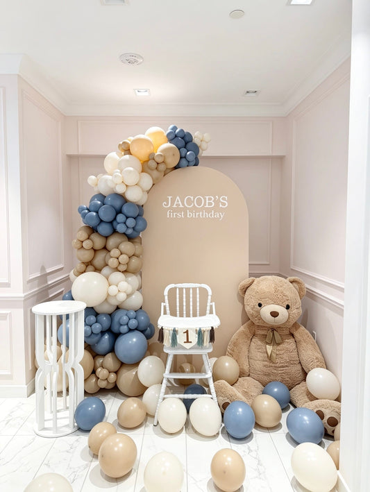 beige full arch backdrop with blue and ivory balloon garland teddy bear and first birthday chair setup