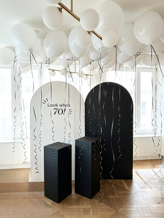 Black and white 70th birthday backdrop with two arch panels, 25 white ceiling helium balloons, black ribbons and pedestal stands.