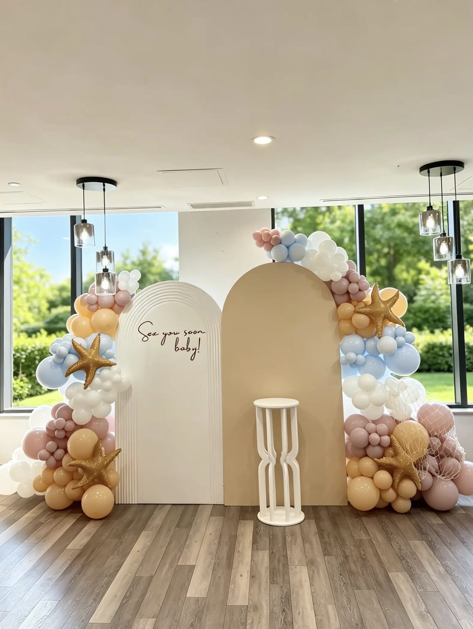 Sea You Soon baby shower backdrop with white ripple and beige arch backdrops, pastel balloon garlands, starfish accents, fishing net decor and a white cake pedestal.
