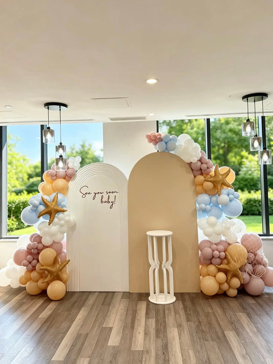 Sea You Soon baby shower backdrop with white ripple and beige arch backdrops, pastel balloon garlands, starfish accents, fishing net decor and a white cake pedestal.