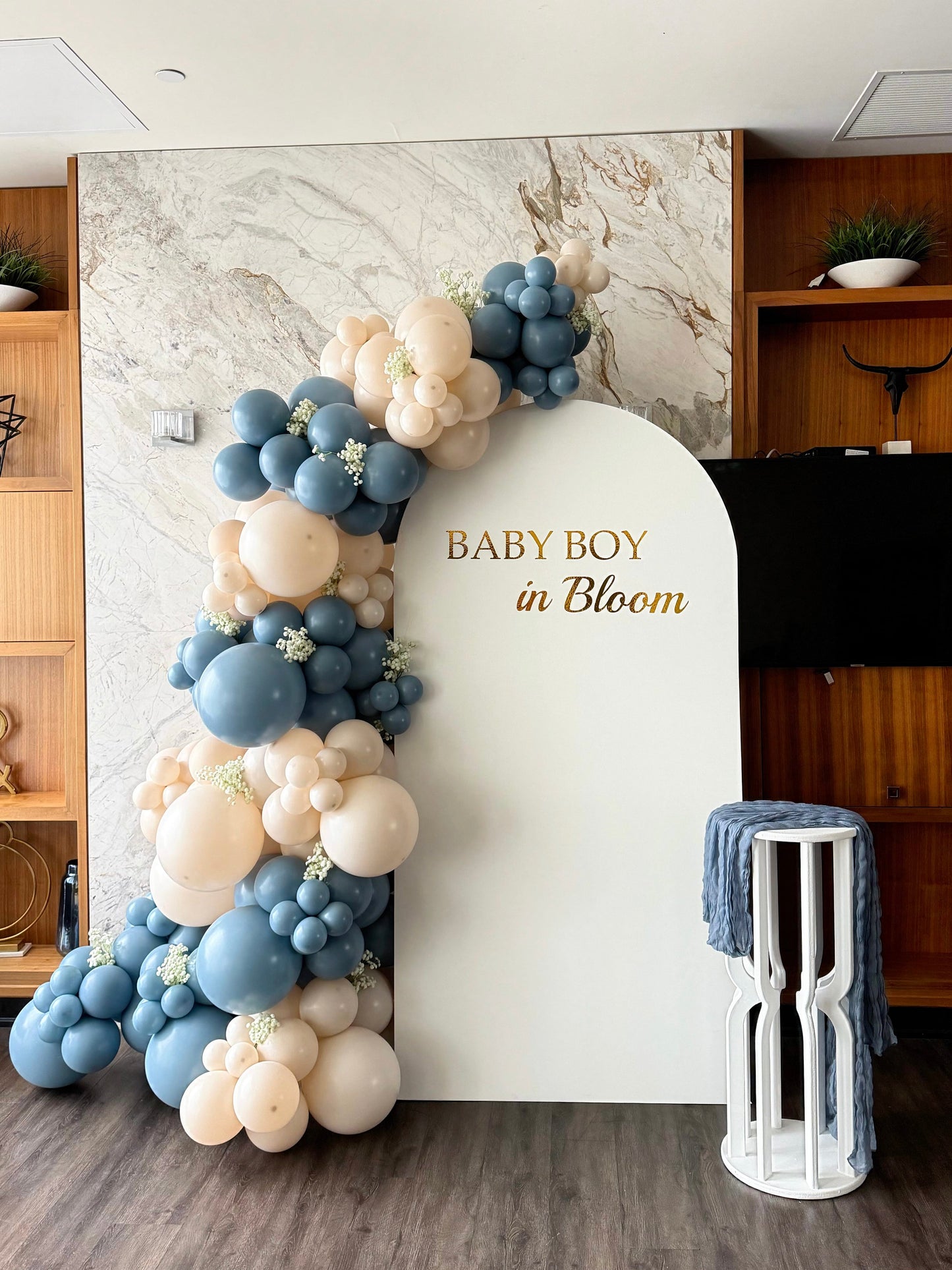 Baby Boy in Bloom backdrop featuring a white arch panel with custom gold lettering, blue and ivory balloon garland, faux baby’s breath accents and a white pedestal stand with blue fabric draping.