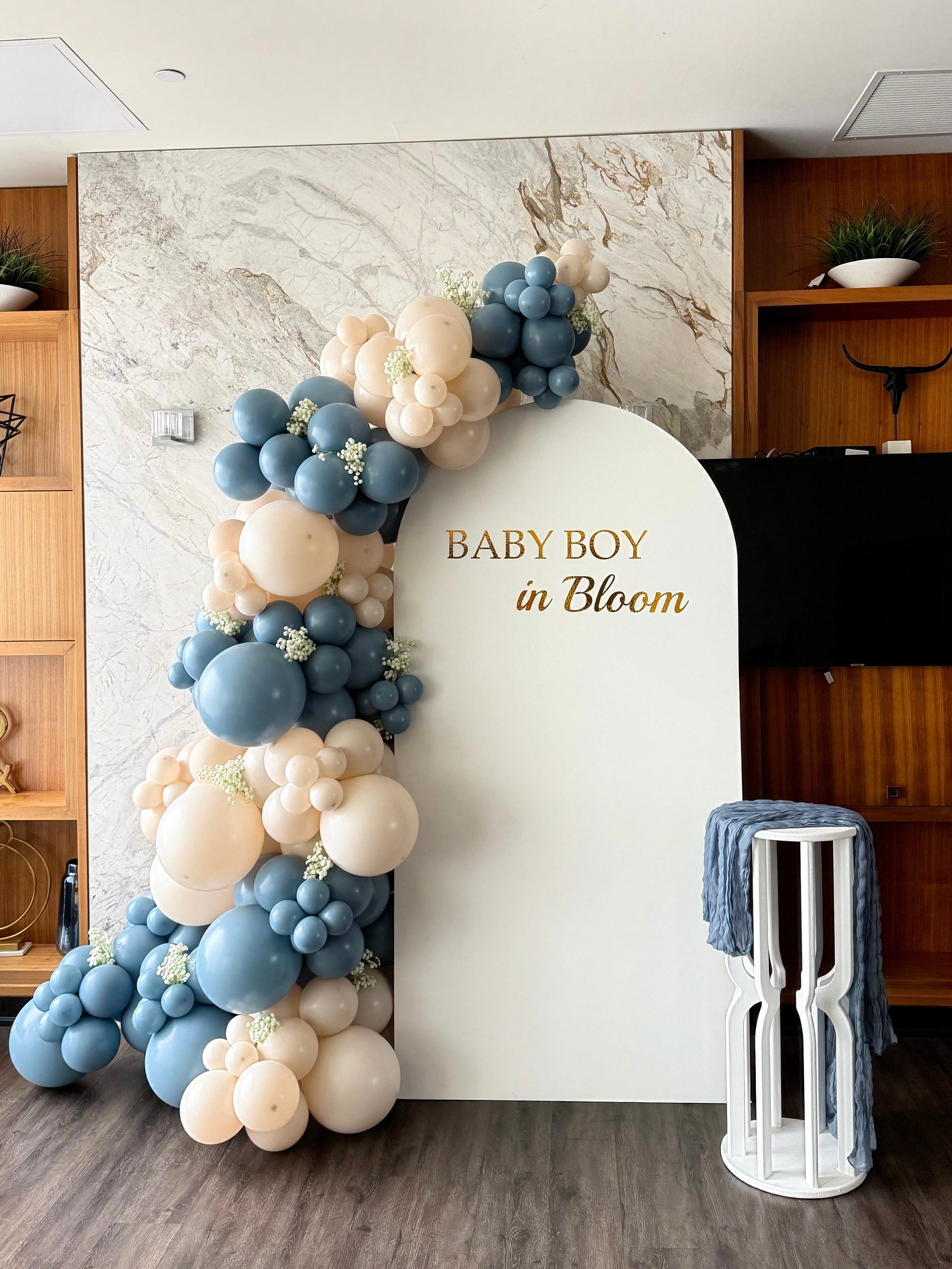 Baby Boy in Bloom backdrop featuring a white arch panel with custom gold lettering, blue and ivory balloon garland, faux baby’s breath accents and a white pedestal stand with blue fabric draping.