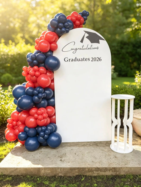 A graduation arch backdrop with a white arched sign reading 'Congratulations Graduates 2026' and a cluster of red, navy, and white balloons on the left, plus a white hourglass decoration on the right, set outdoors on a stone base with greenery in the background.