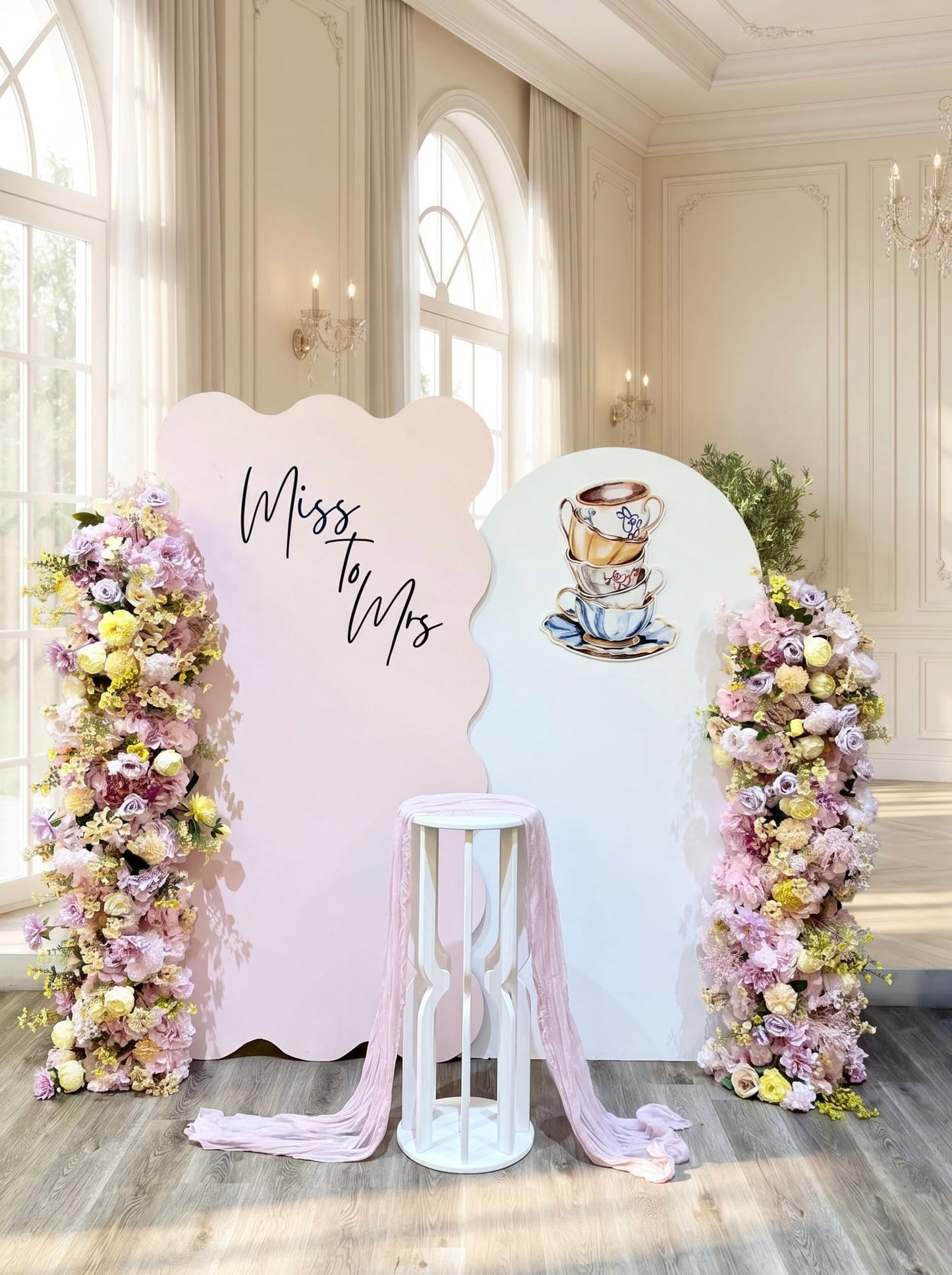 Tea party bridal shower backdrop featuring pastel pink and white wavy panels, custom Miss to Mrs lettering, teacup artwork, luxury faux floral arrangements and a white pedestal stand.