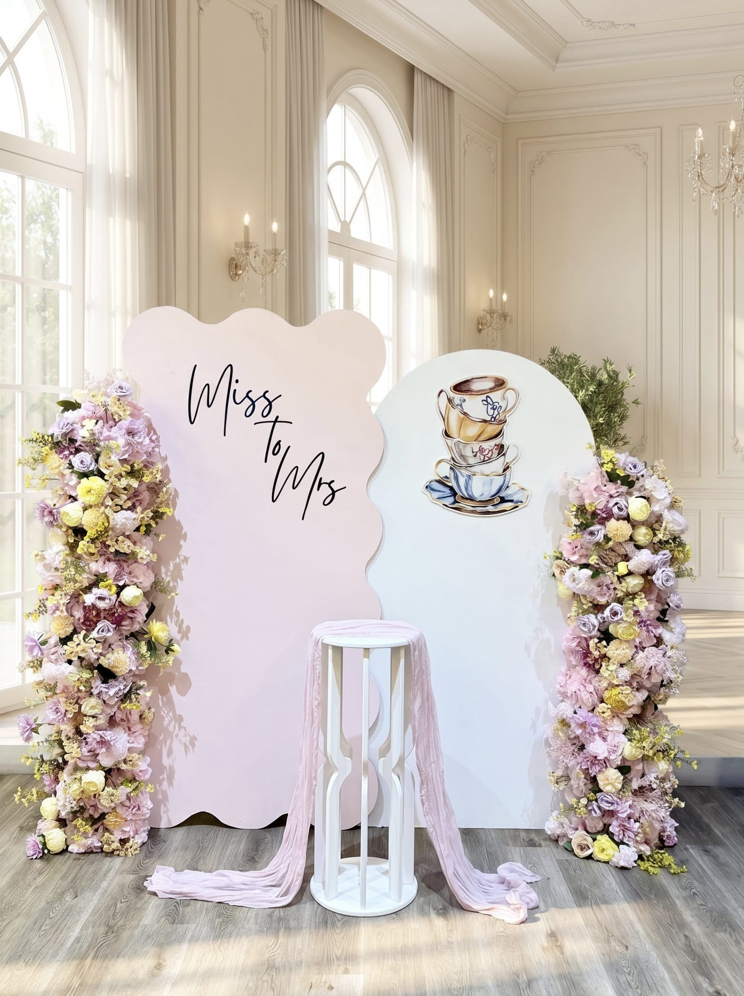 Tea party bridal shower backdrop featuring pastel pink and white wavy panels, custom Miss to Mrs lettering, teacup artwork, luxury faux floral arrangements and a white pedestal stand.