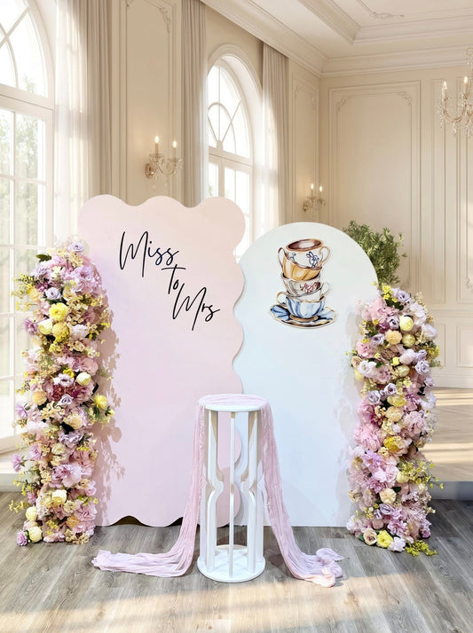 Tea party bridal shower backdrop featuring pastel pink and white wavy panels, custom Miss to Mrs lettering, teacup artwork, luxury faux floral arrangements and a white pedestal stand.