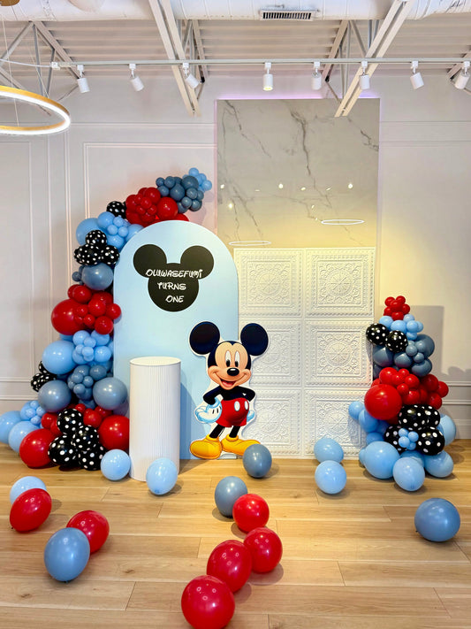 Mickey Mouse first birthday setup with light blue arch and white textured backdrop, red blue black and white balloons, character cutout and pedestal