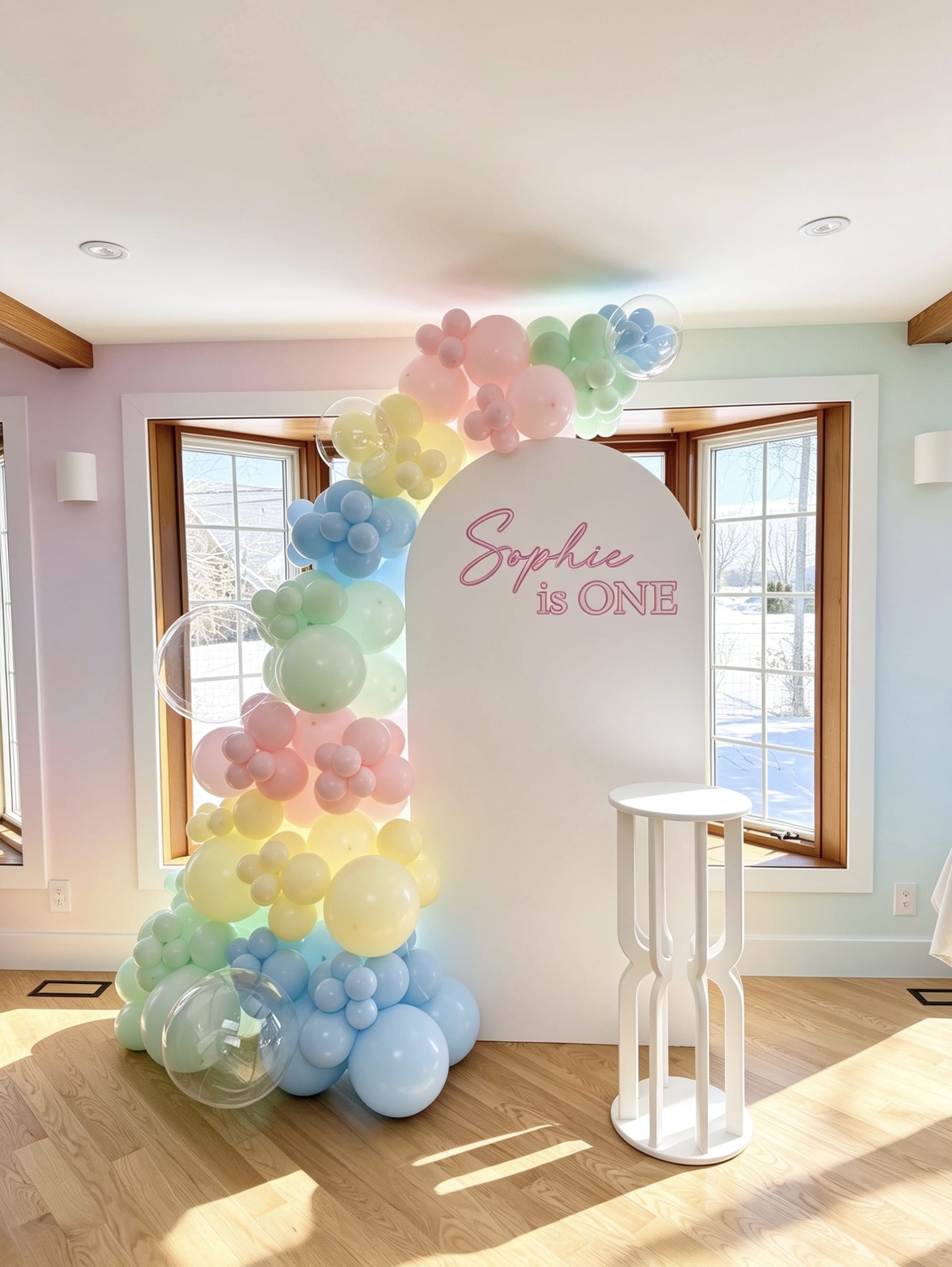 pastel rainbow first birthday setup with white full arch backdrop pastel balloons and cake table