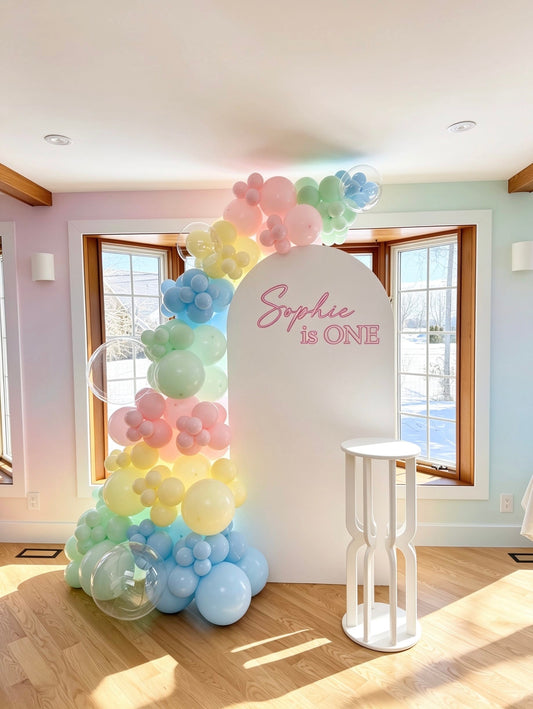 pastel rainbow first birthday setup with white full arch backdrop pastel balloons and cake table