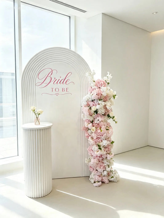 White ripple arch bridal shower backdrop with Bride to Be sign, pink and white floral arrangement and white ribbed pedestal