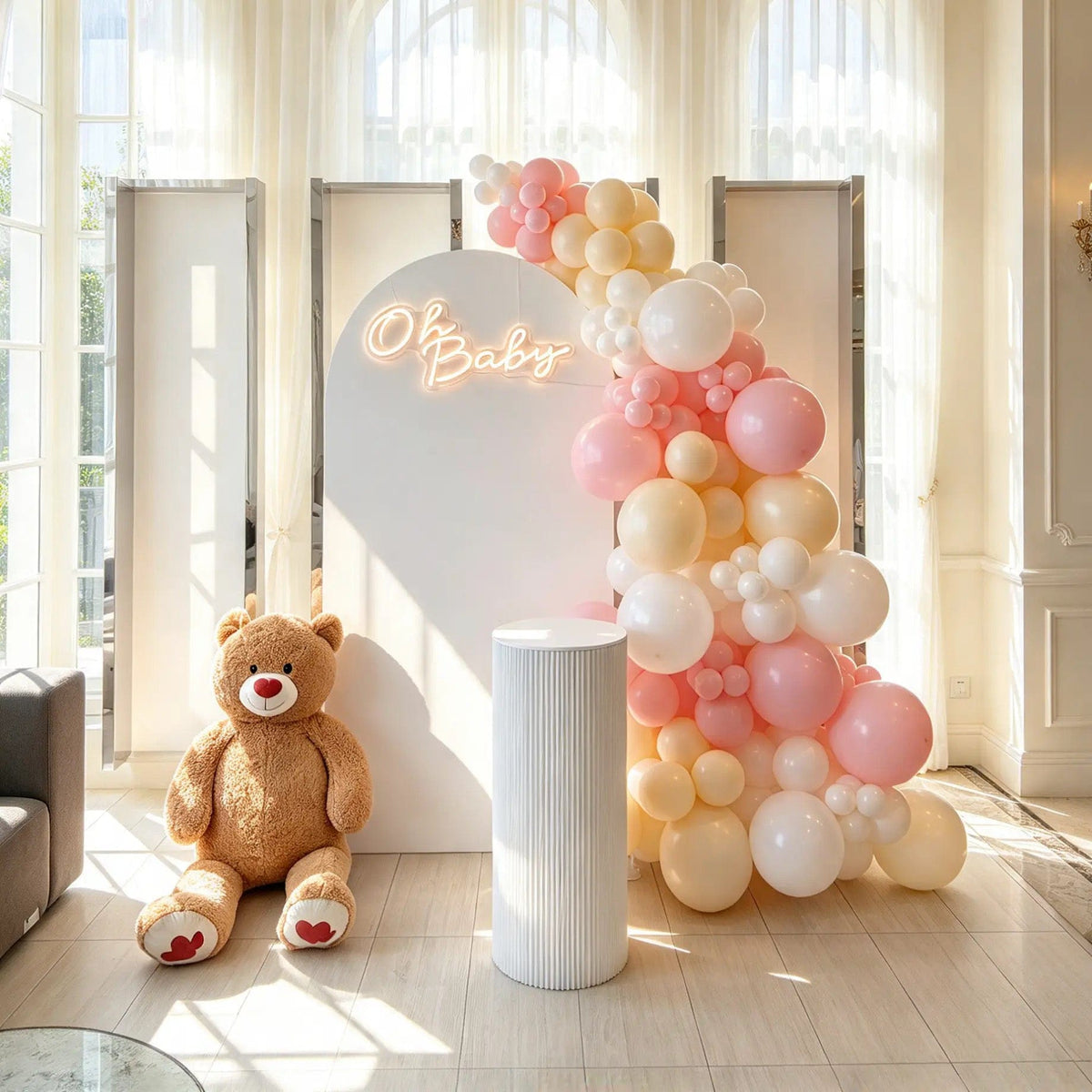 White baby shower arch with a glowing “Oh Baby” sign, pink, cream, and white balloons, a large teddy bear, and a white cake pedestal.