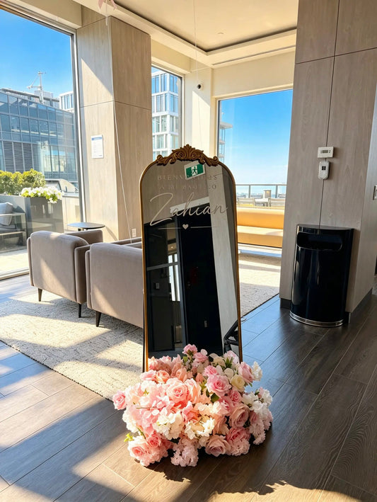 Ornate gold mirror welcome sign with personalized baptism lettering and a pink and white floral arrangement at the base.