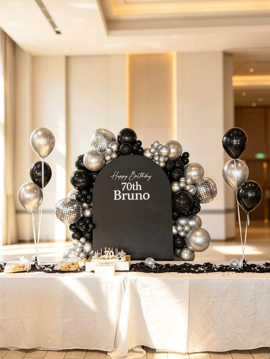 Black milestone birthday backdrop featuring a custom arch panel with black, silver and chrome balloon garland, disco balloon accents and two matching three-balloon centerpieces.