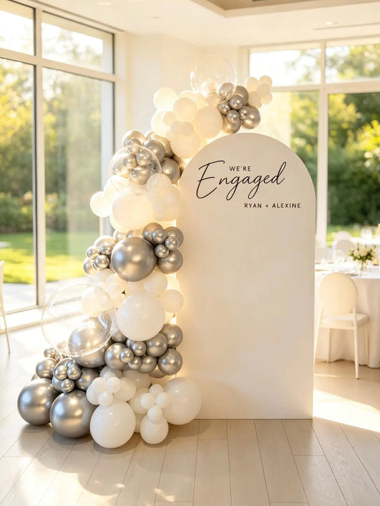Engagement backdrop with white arch, personalized names, white and metallic silver balloon garland and clear bubble balloons.