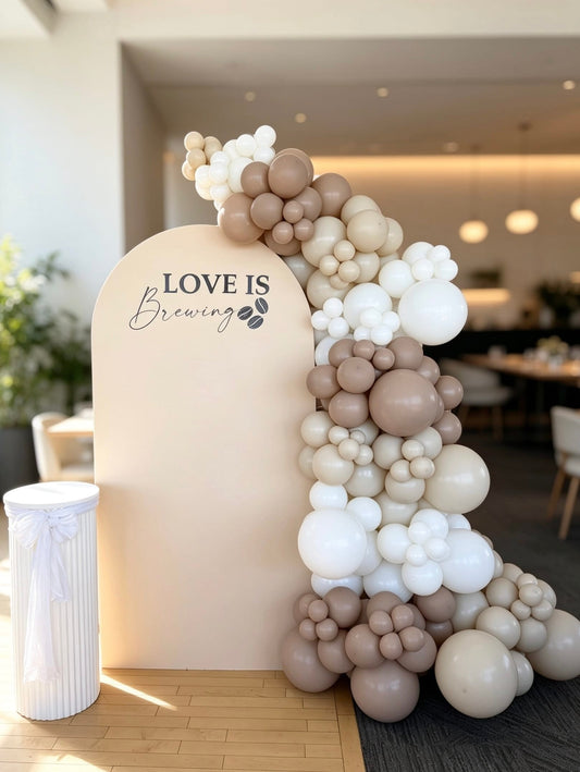 Neutral Love Is Brewing bridal shower backdrop with beige and ivory balloon garland, custom arch backdrop and white pedestal stand.