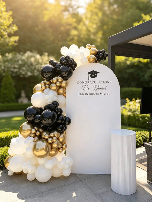 Graduation backdrop with personalized name, black, gold and white organic balloon garland and white ribbed cake table.