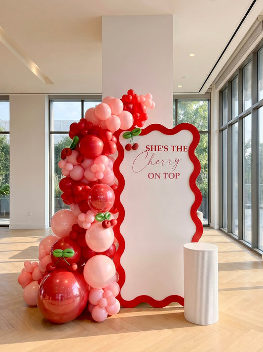 Cherry-themed birthday backdrop featuring a white wavy panel with red border, custom lettering, pink and red balloon garland, cherry accents, and a white ribbed pedestal stand.
