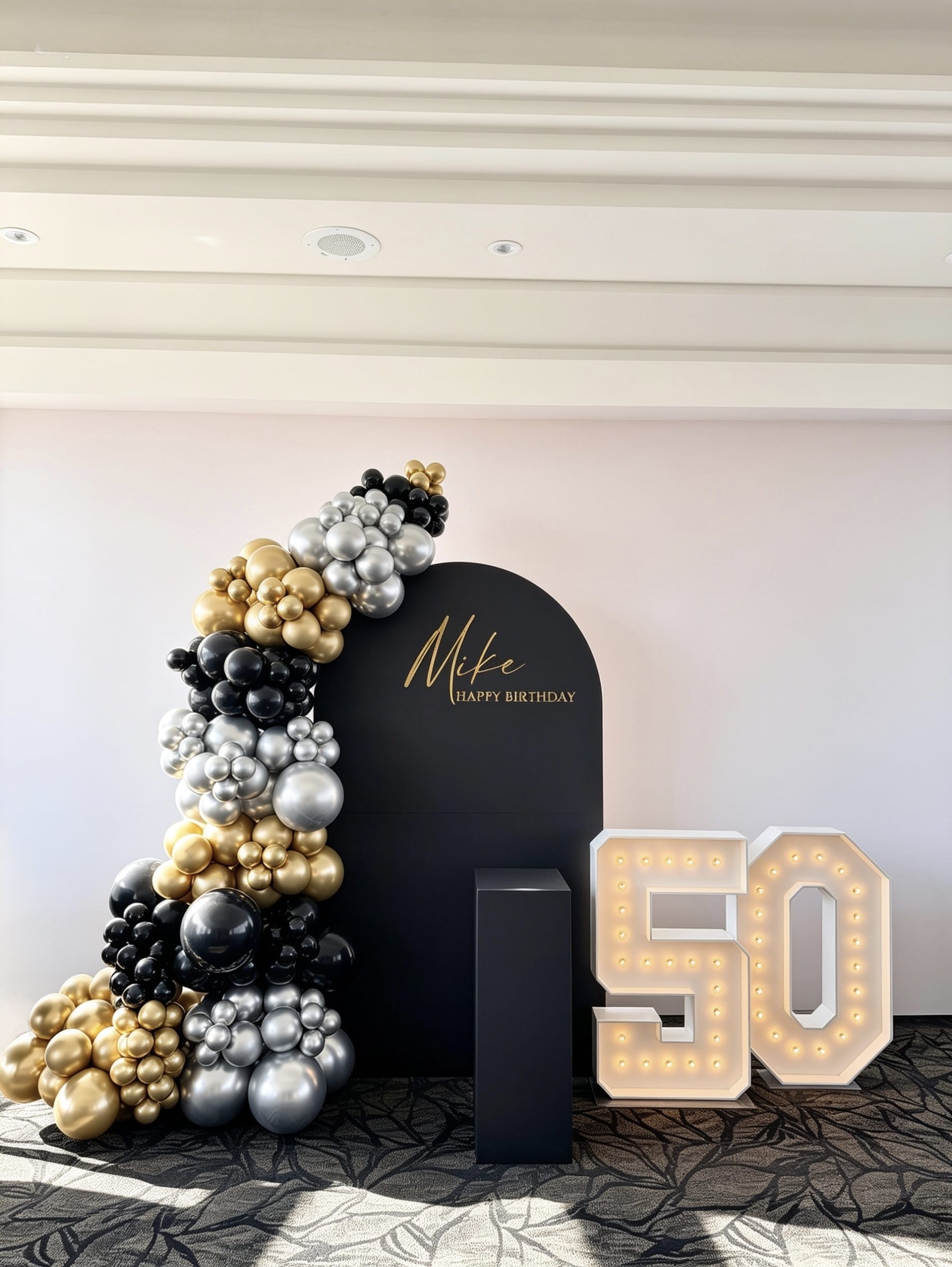 Black arch backdrop with black, gold and silver balloon garland and illuminated 50 marquee numbers for a 50th birthday celebration